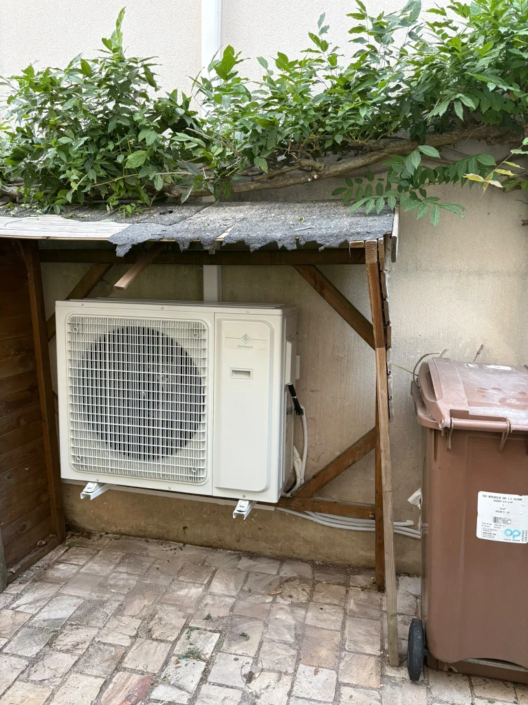 Groupe extérieur de climatisation installé sous un auvent en bois dans une cour pavée, climaticien Île-de-France