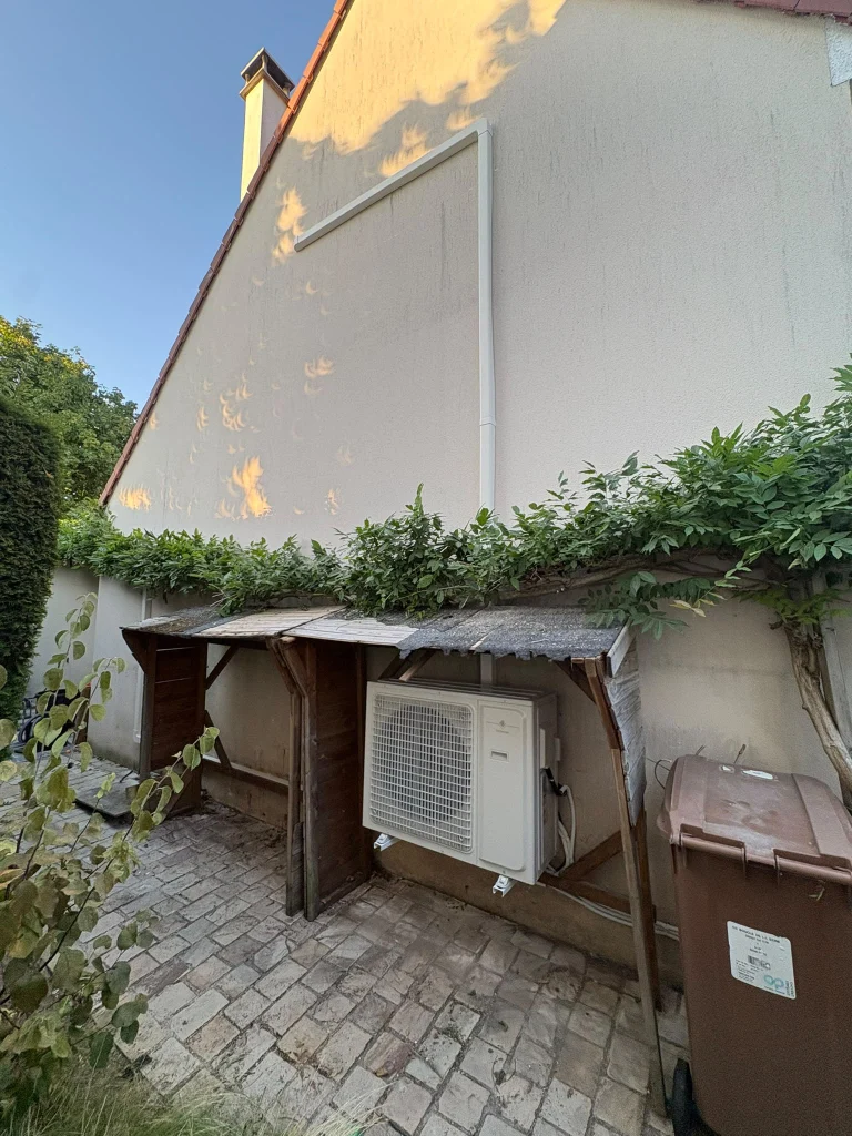 Groupe extérieur de climatisation installé sous un auvent en bois dans une cour pavée, climaticien Île-de-France