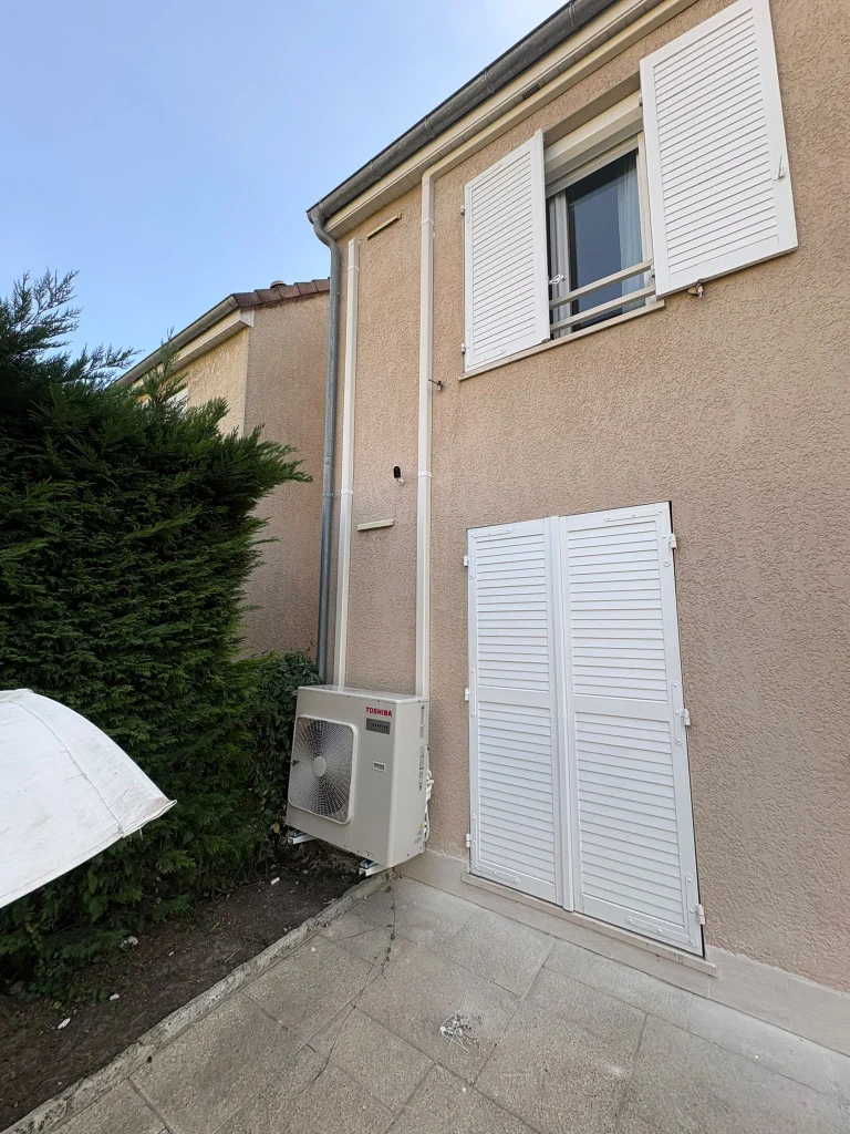 Groupe extérieur de climatisation au sol le long d'un pavillon aux volets blancs, climaticien Île-de-France