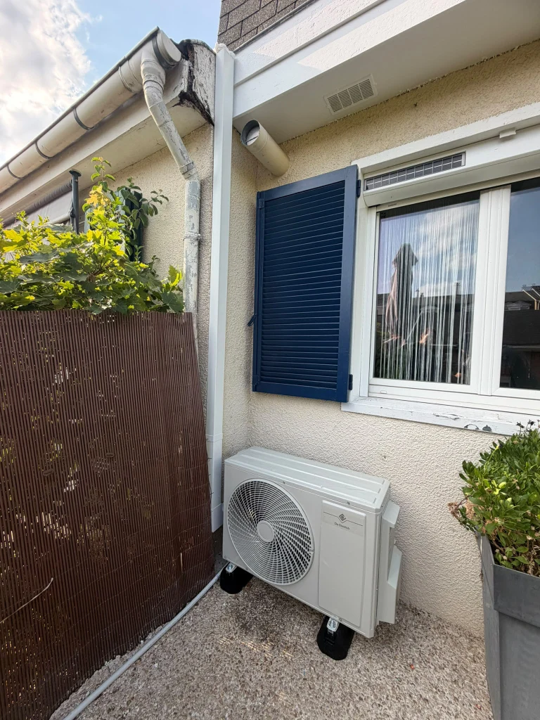 Groupe extérieur de climatisation au sol devant une maison aux volets bleus et clôture bois, climaticien Île-de-France