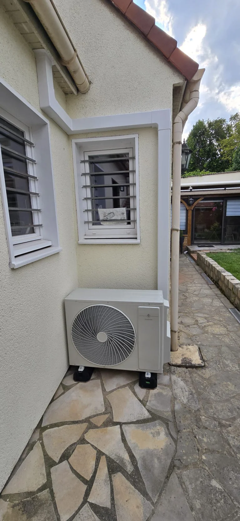 Groupe extérieur de climatisation au sol sur une allée en pierre le long d'un pavillon crème, climaticien Île-de-France