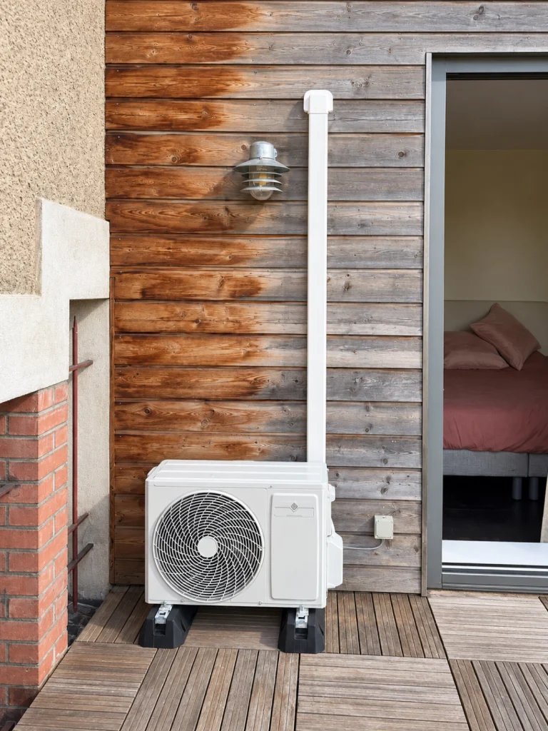 Groupe extérieur de climatisation réversible sur plots devant un bardage bois, goulotte remontant vers la chambre, installation de climatisation Île-de-France
